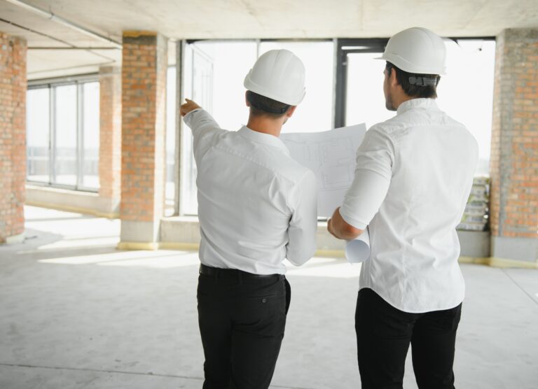 A front view of two smart architects with white helmets reviewing blueprints at a construction site