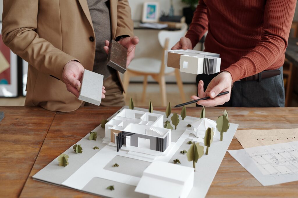 Young architect pointing at model of new house and his mature colleague choosing suitable block
